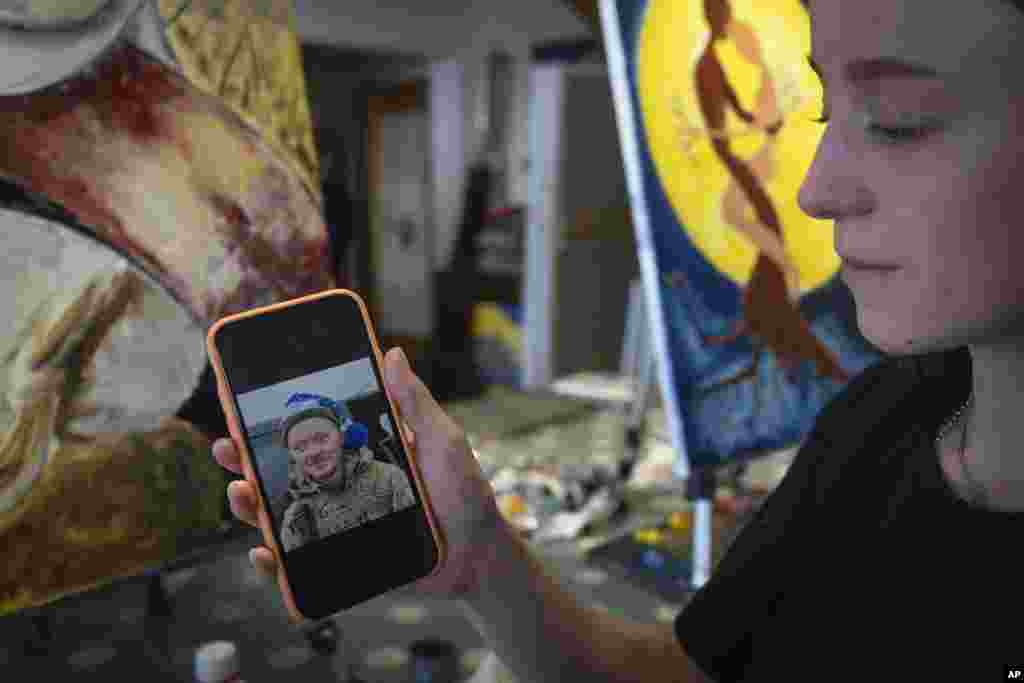 Olesia Skalska arată o fotografie a soțului ei, Roman Skalski, care a fost ucis într-o luptă cu trupele rusești.&nbsp; Ea a venit la studioul de artă cu Irina Farion, pe care a întâlnit-o la un cimitir, un loc în care văduvele ucrainene găsesc consolare una în compania celeilalte și formează prietenii bazate pe durerea comună. În vârstă de 26 de ani, Olesia s-a înrolat voluntar în armată, deși nu avea experiență anterioară de luptă. Cuplul trebuia să sărbătorească prima aniversare a nunții în iunie. Dar, în schimb, Skalska lucrează la un tablou, singurul loc unde pot fi din nou împreună. &bdquo;Un bărbat duce o fată pe un câmp de grâu care a fost tăiat și îmi imaginez că o poartă astfel încât să nu-și înțepe picioarele&rdquo;, spune Skalska, în vârstă de 24 de ani, descriindu-și opera de artă.