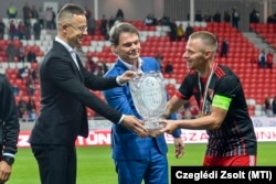 Szijjártó Péter adja át a másodosztályú bajnoki serleget a diplomata-útlevéllel is rendelkező Dzsudzsák Balázsnak Debrecenben, 2021. május 16-án. Szijjártó mellett világoskék zakóban a fideszes Bánki Erik áll, a parlament gazdasági bizottságának elnöke.